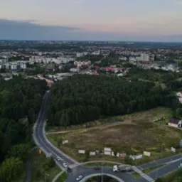 Panorama miasta z lotu ptaka o zmierzchu, widoczne bloki mieszkalne, las, drogi z ruchem samochodowym i ronda.