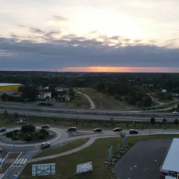 Zdjęcie z drona przedstawiające rondo z samochodami w ruchu, drogę szybkiego ruchu, żółty budynek magazynowy w tle oraz zachodzące słońce za linią lasu.