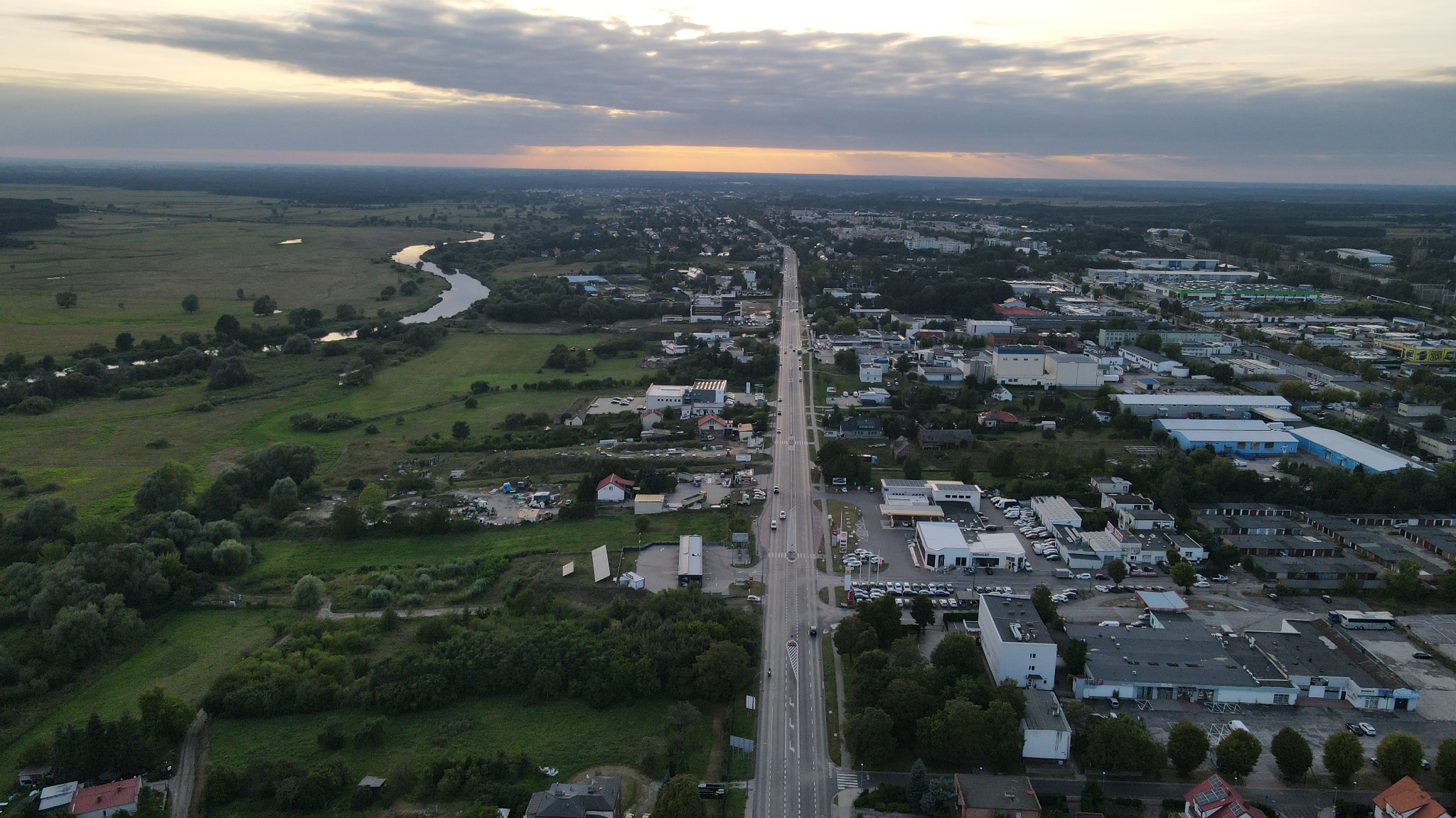 Panoramiczny widok z lotu ptaka na miasto z długą prostą drogą, rzeką wijącą się po lewej stronie i zachodzącym słońcem na horyzoncie.