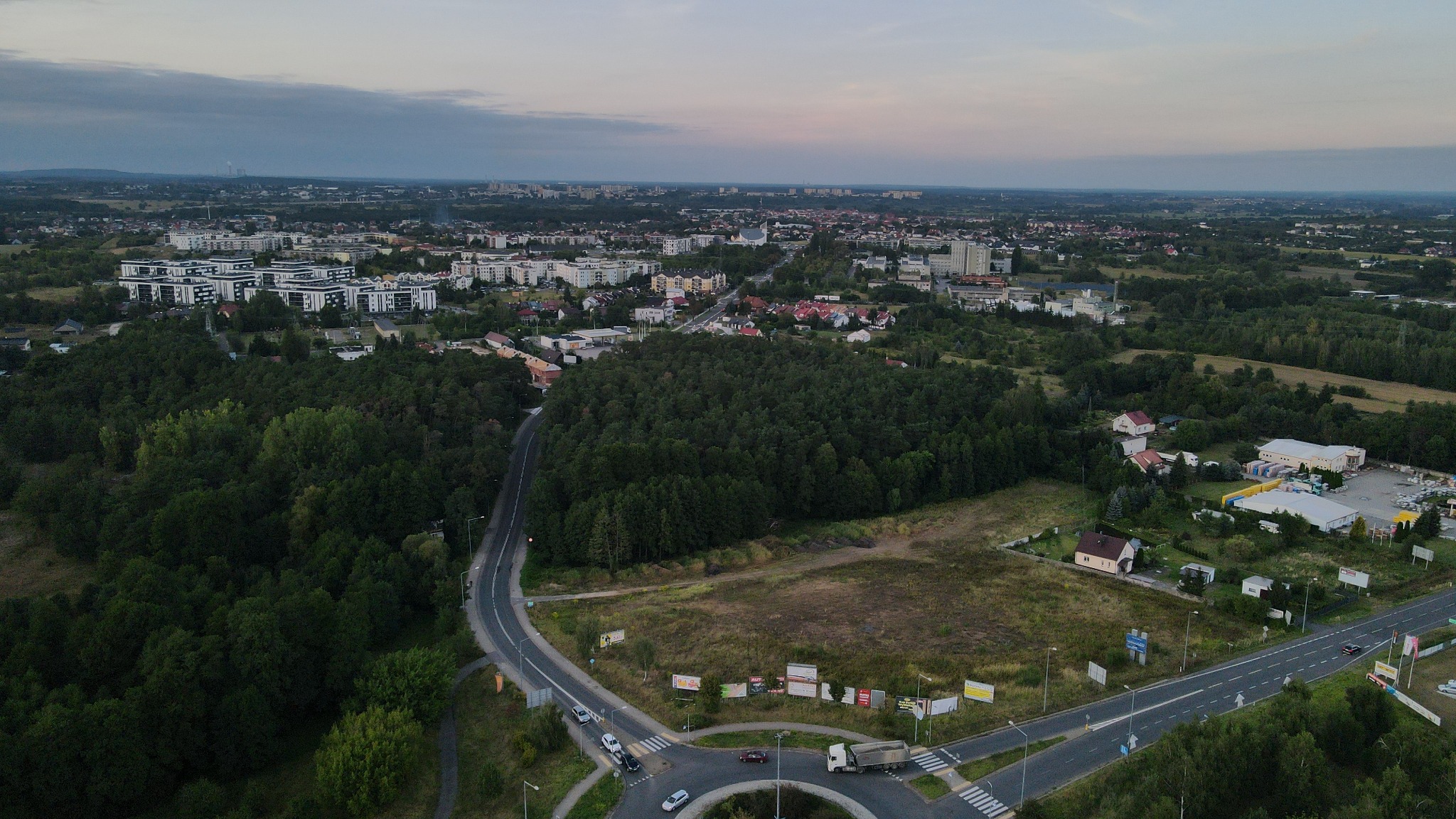 Panorama miasta z lotu ptaka o zmierzchu, widoczne bloki mieszkalne, las, drogi z ruchem samochodowym i ronda.