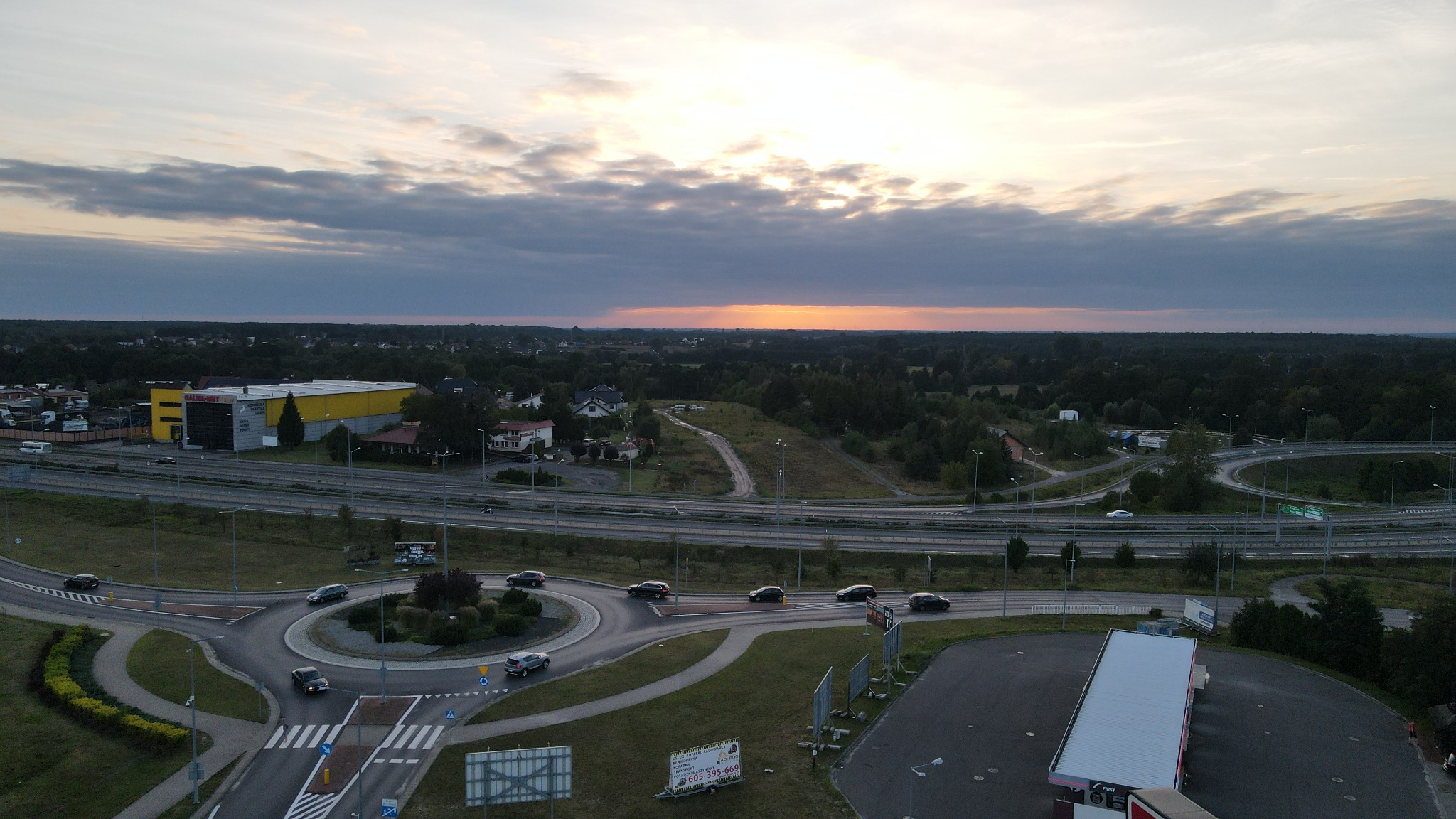 Zdjęcie z drona przedstawiające rondo z samochodami w ruchu, drogę szybkiego ruchu, żółty budynek magazynowy w tle oraz zachodzące słońce za linią lasu.