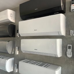 SP EKO GROUP SPÓŁKA Z OGRANICZONĄ ODPOWIEDZIALNOŚCIĄ - Ekspozycja różnych modeli klimatyzatorów marki Gree w kolorach białym, srebrnym i czarnym, wraz z pilotami, zamontowanych na szarej ścianie.