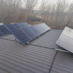 Panele fotowoltaiczne zamontowane na dachu z blachy trapezowej, widoczne kable prowadzące po dachu, drzewa bez liści w tle.