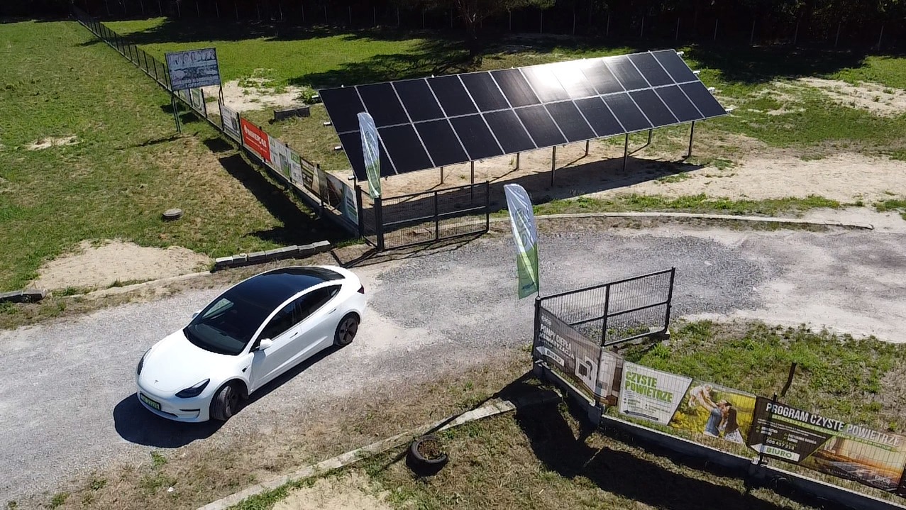 Widok z góry na białą Teslę zaparkowaną przed konstrukcją z paneli słonecznych na piaszczystym terenie z ogrodzeniem reklamowym w tle.