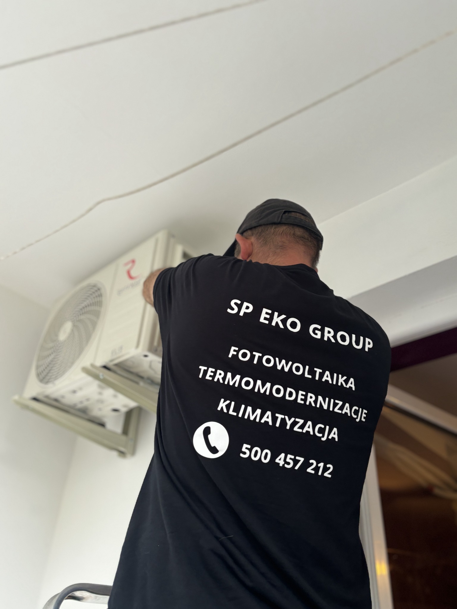 Instalator w czarnej koszulce firmowej SP EKO GROUP montuje klimatyzator na ścianie wewnątrz pomieszczenia.