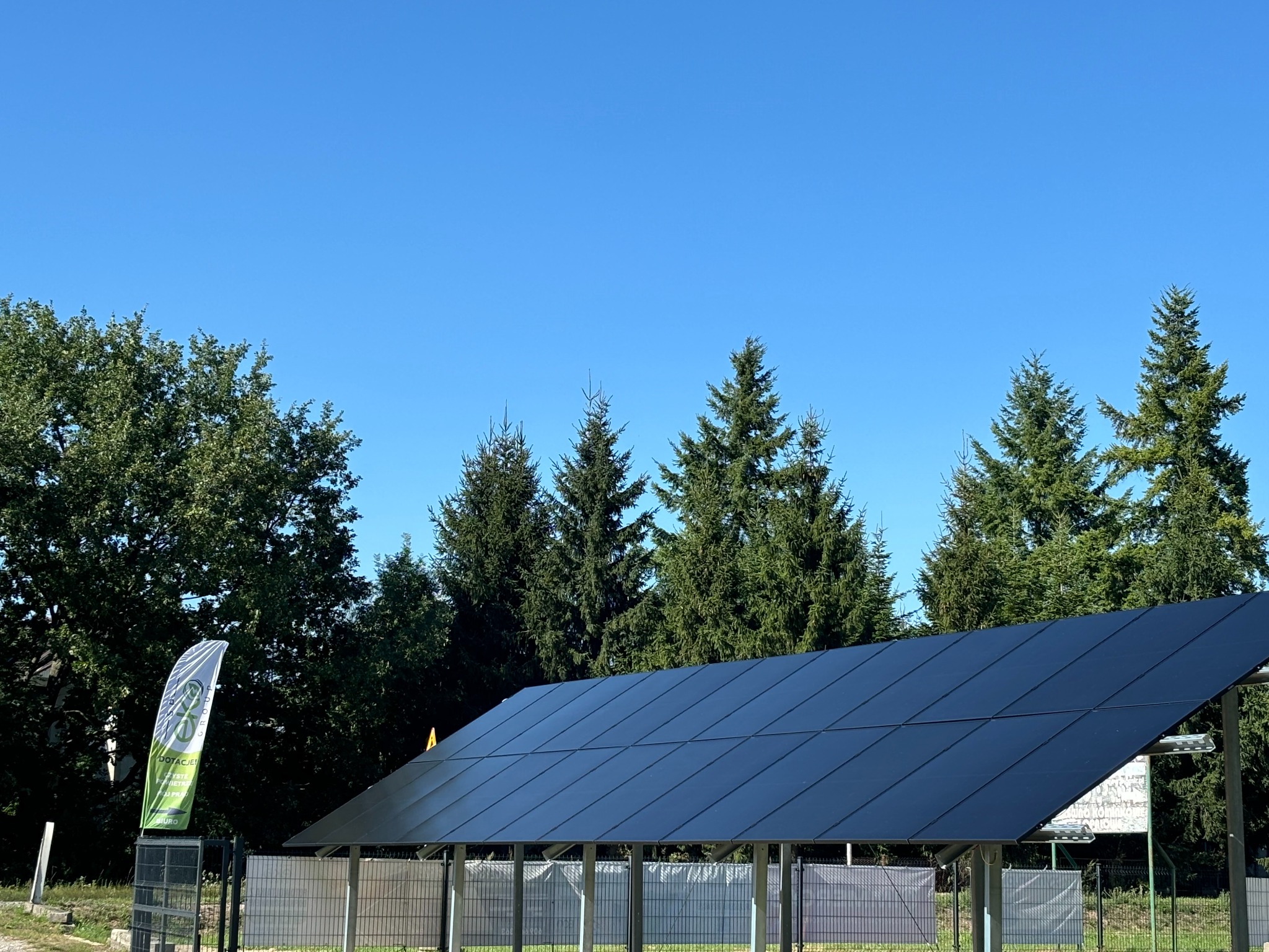 Czarny panel fotowoltaiczny zamontowany jako zadaszenie, widoczny na tle zielonych drzew i błękitnego nieba, z flagą reklamową po lewej stronie.
