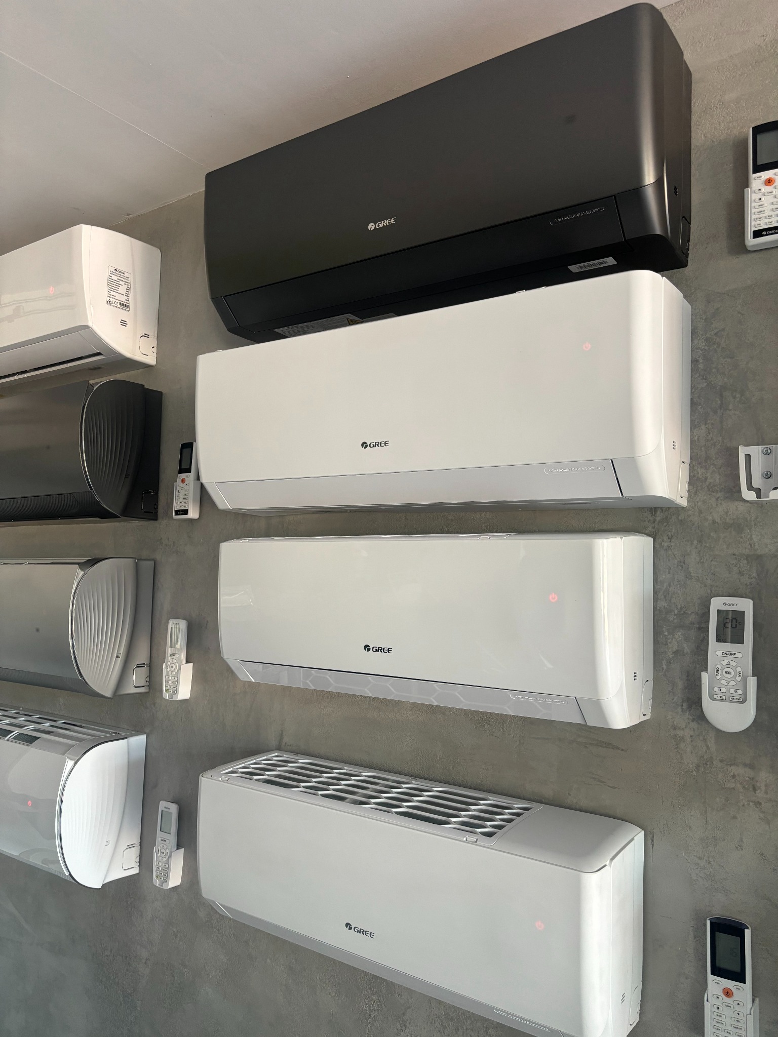 Ekspozycja różnych modeli klimatyzatorów marki Gree w kolorach białym, srebrnym i czarnym, wraz z pilotami, zamontowanych na szarej ścianie.
