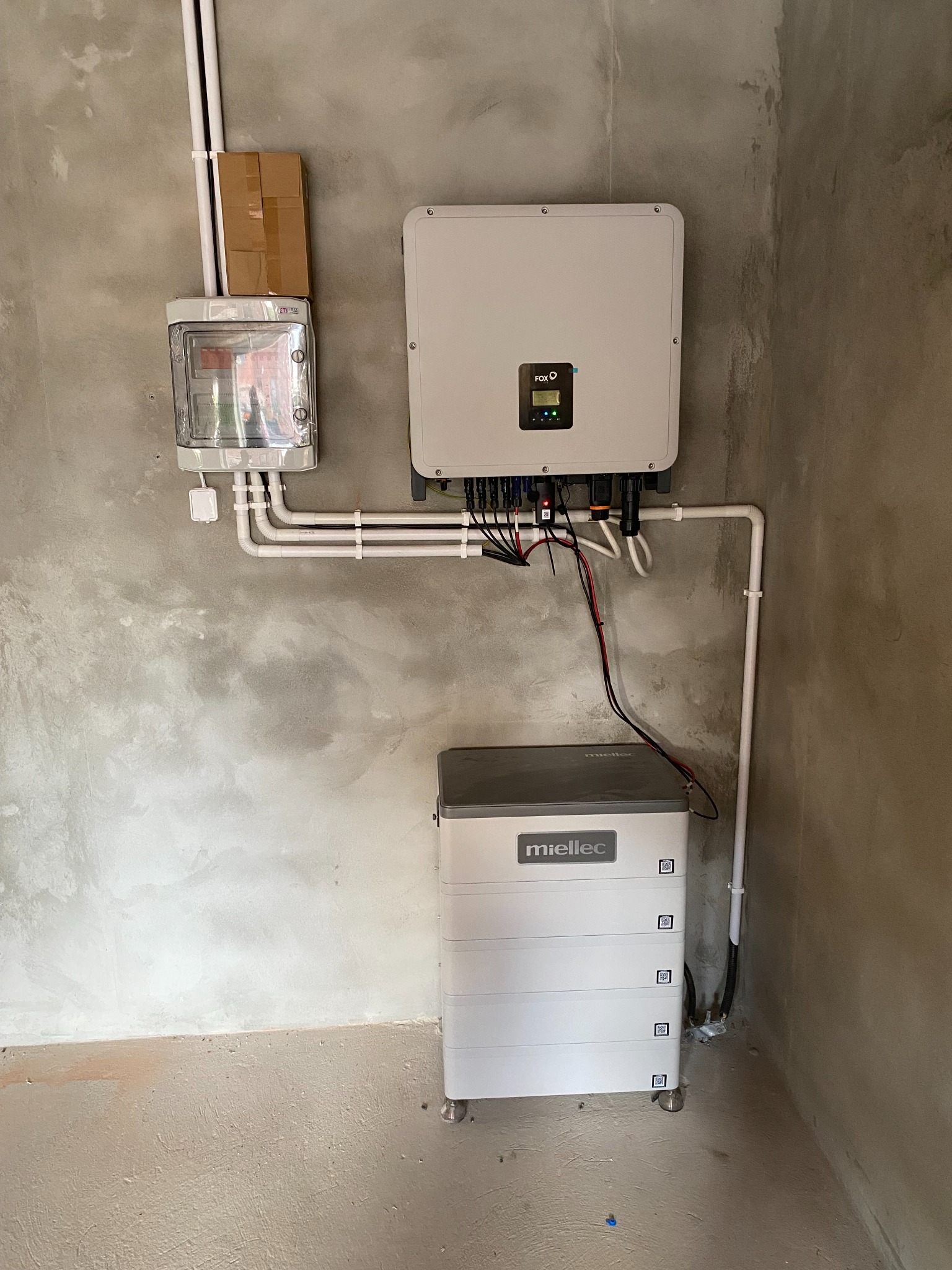 Zamontowany system fotowoltaiczny w surowym pomieszczeniu: falownik FOX i bateria Mieliec, widoczne okablowanie i licznik energii.