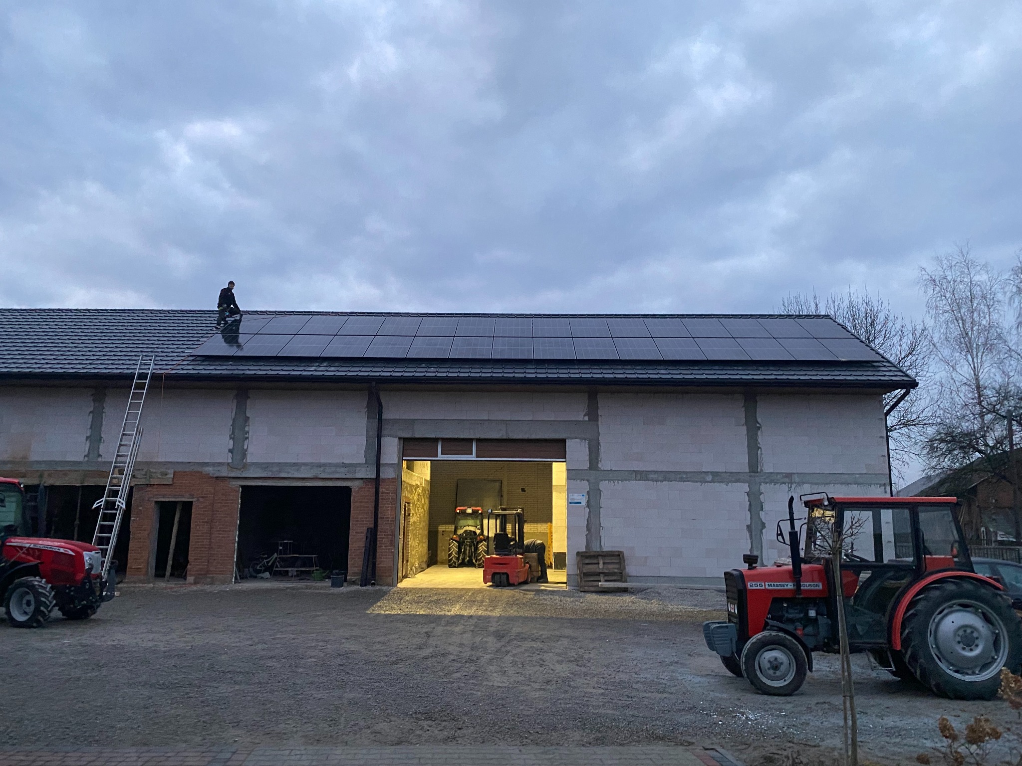 Montaż paneli słonecznych na dachu budynku gospodarczego; w tle widoczne traktory i wózek widłowy.