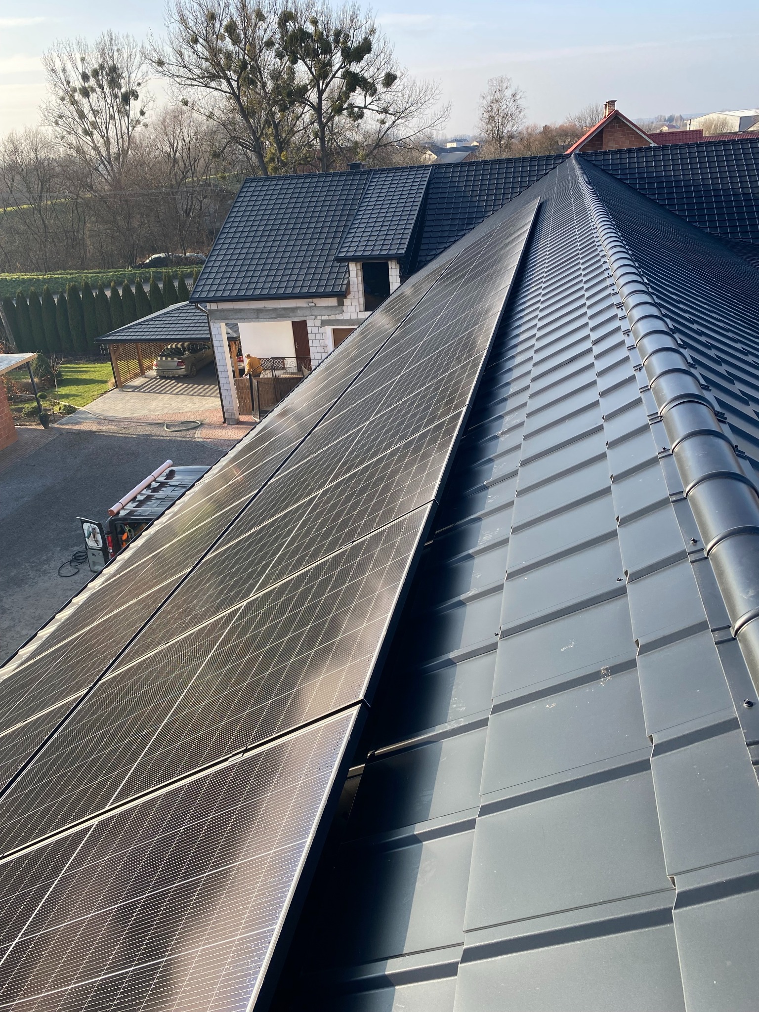 Częściowy widok dachu z panelami fotowoltaicznymi w kolorze czarnym, obok blaszana dachówka w kolorze grafitowym, w tle dom z podjazdem i zielenią.