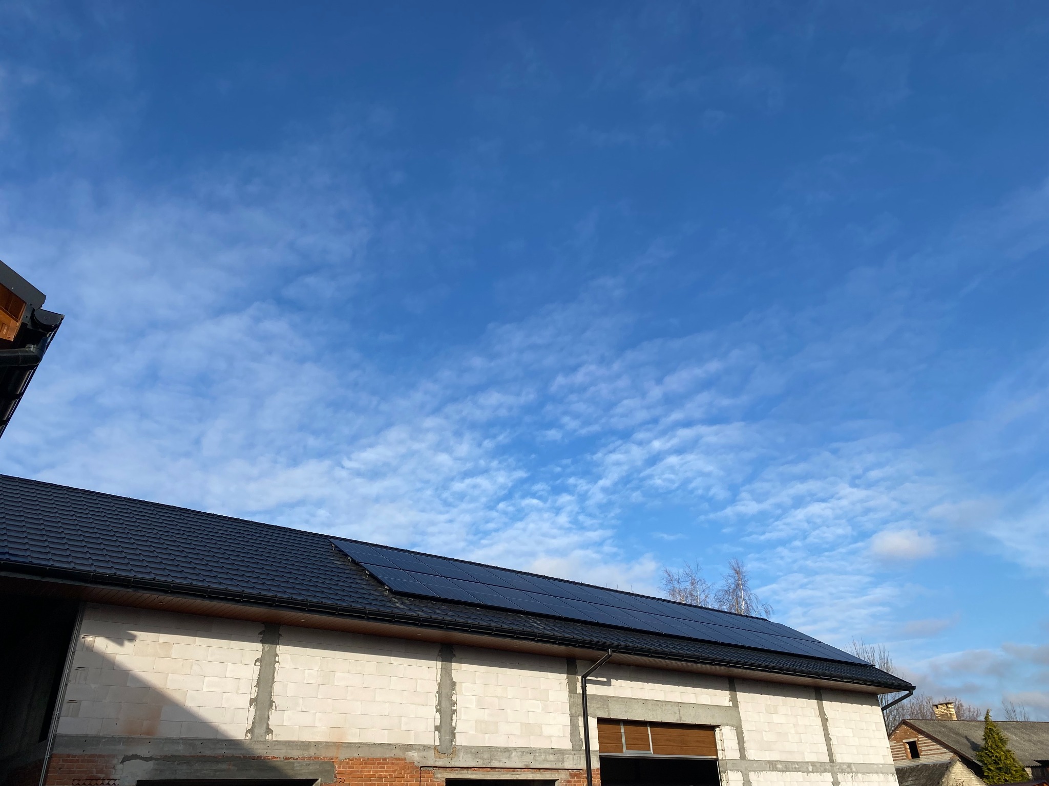 Panele fotowoltaiczne zamontowane na dachu budynku w trakcie budowy, widoczne białe bloczki i czerwona cegła, błękitne niebo z białymi obłokami w tle.