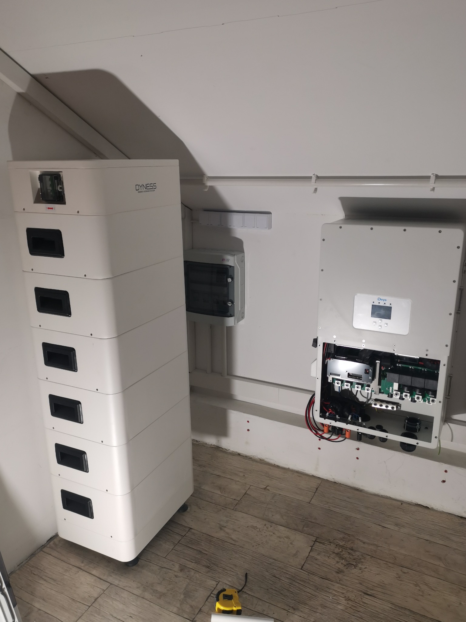 Biały moduł bateryjny Dyness ustawiony obok otwartego falownika Deye z widocznymi przewodami, zamontowanego na białej ścianie w pomieszczeniu z drewnianą podłogą, z żółtą miarką leżącą na podłodze.