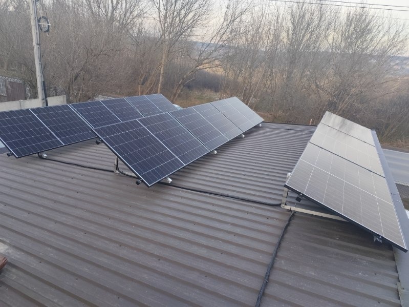 Panele fotowoltaiczne zamontowane na dachu z blachy trapezowej, widoczne kable prowadzące po dachu, drzewa bez liści w tle.