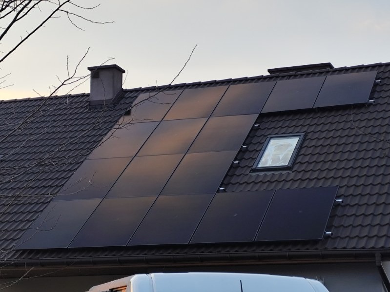 Ciemne panele fotowoltaiczne na spadzistym dachu z dachówki ceramicznej, obok okna dachowego i komina; częściowo widoczny biały samochód na dole kadru.