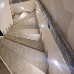 Usługi budowlane płytki - Schody wyłożone płytkami imitującymi kamień, z zamontowanymi punktowymi lampami w ścianie, z zabezpieczonymi elementami do montażu poręczy.