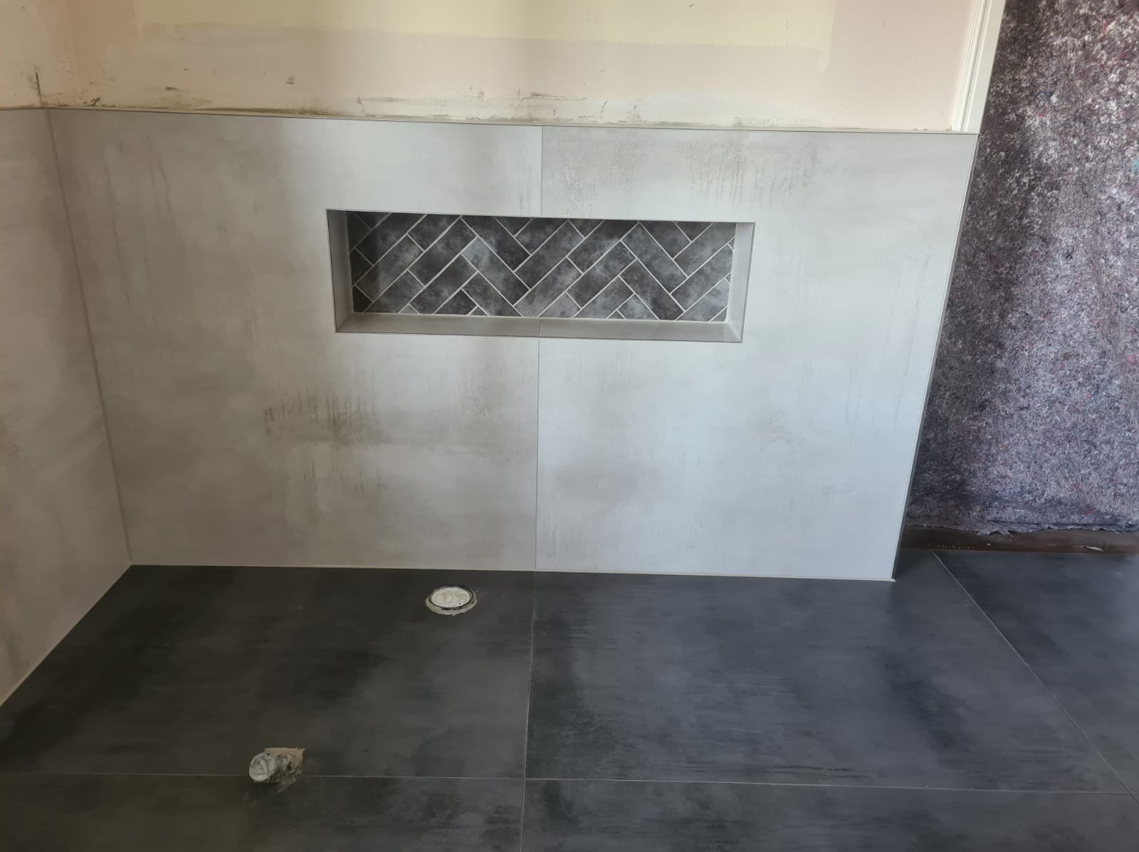 Świeżo wykafelkowana ściana prysznica z wnęką wyłożoną szarą mozaiką w jodełkę, ciemnoszara podłoga z odpływem, widoczne niedoskonałości fugowania.