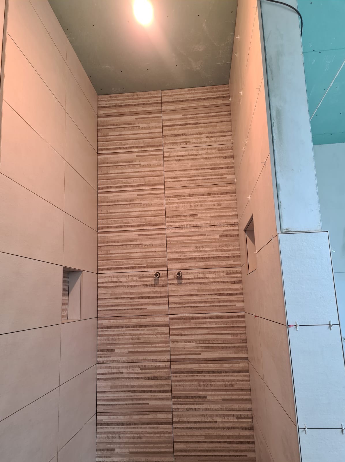 Ściana łazienki wyłożona beżowymi płytkami w dwóch wzorach: gładkie, duże płytki i mozaika z poziomych pasków imitujących drewno, widoczne otwory na instalacje i krzyżyki dystansowe.