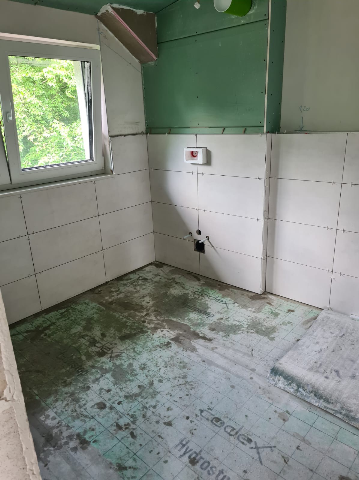 Łazienka w trakcie prac: białe płytki ścienne ułożone do połowy wysokości pomieszczenia, widoczne otwory na instalacje, podłoga pokryta hydroizolacją z widocznymi zabrudzeniami, okno z widokiem...