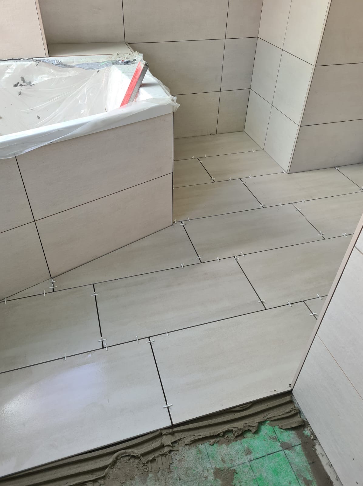 Układanie beżowych płytek wielkoformatowych na podłodze i ścianach łazienki, widoczne krzyżyki dystansowe, wanna zabezpieczona folią.