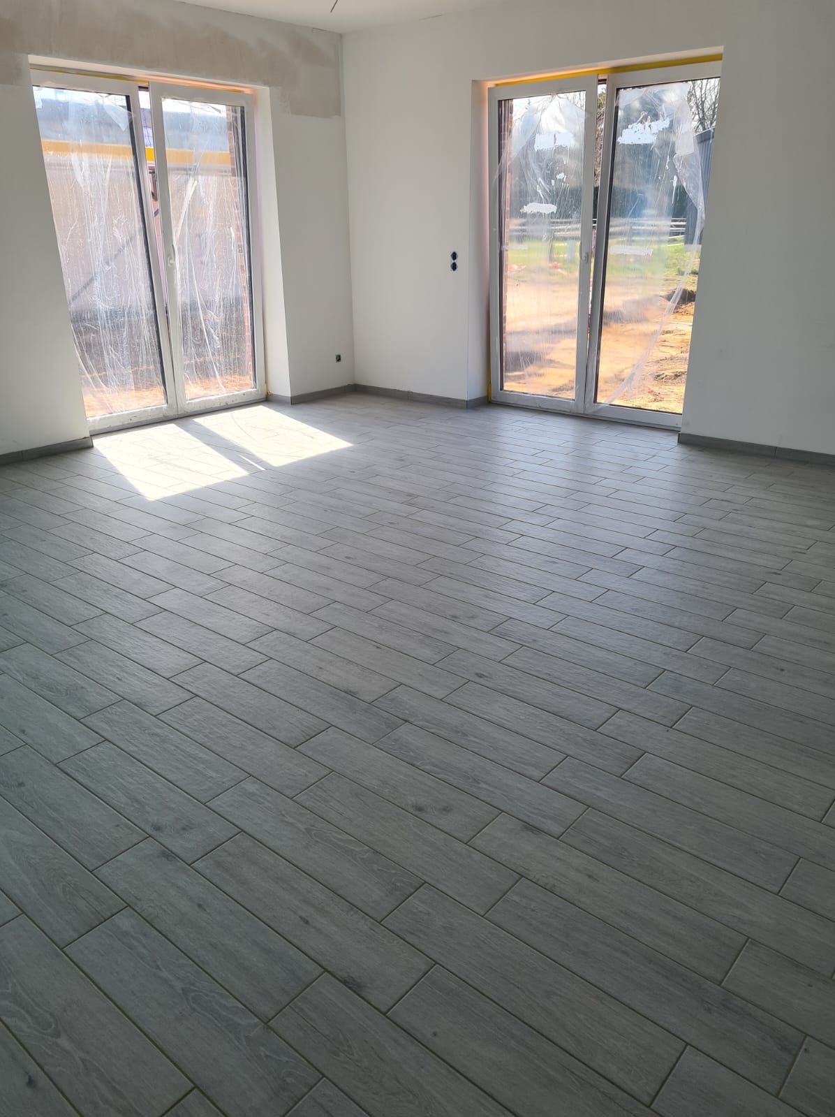 Jasnoszara podłoga z płytek imitujących drewno ułożona w nowym, pustym pomieszczeniu z dwoma oknami zabezpieczonymi folią ochronną.
