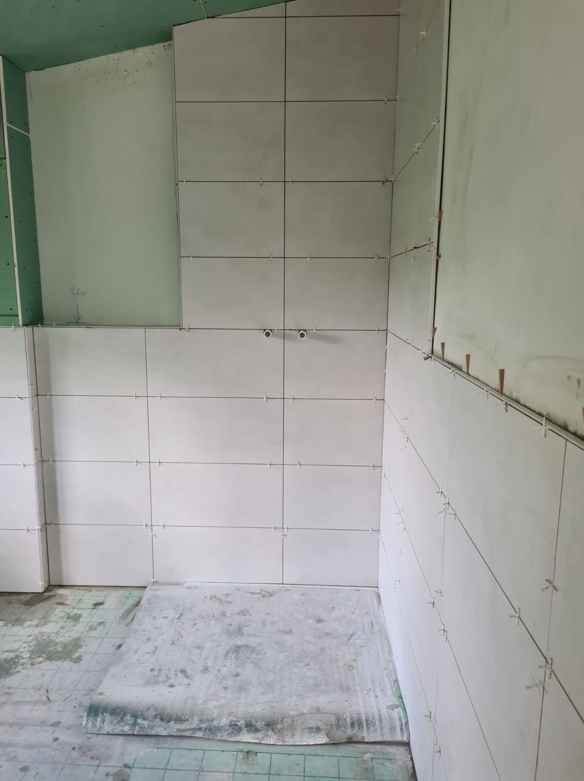 Białe płytki ceramiczne układane na ścianach łazienki, widoczne krzyżyki dystansowe, fragment zielonej płyty kartonowo-gipsowej na ścianie, zabezpieczona folią podłoga.