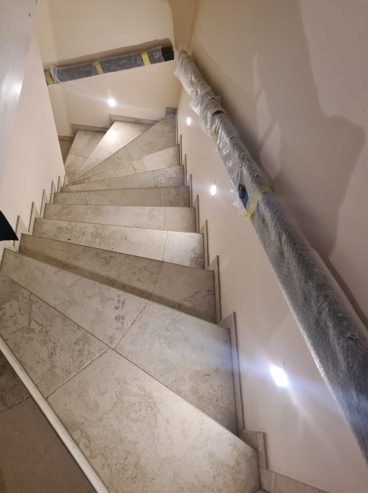 Schody wyłożone płytkami imitującymi kamień, z zamontowanymi punktowymi lampami w ścianie, z zabezpieczonymi elementami do montażu poręczy.