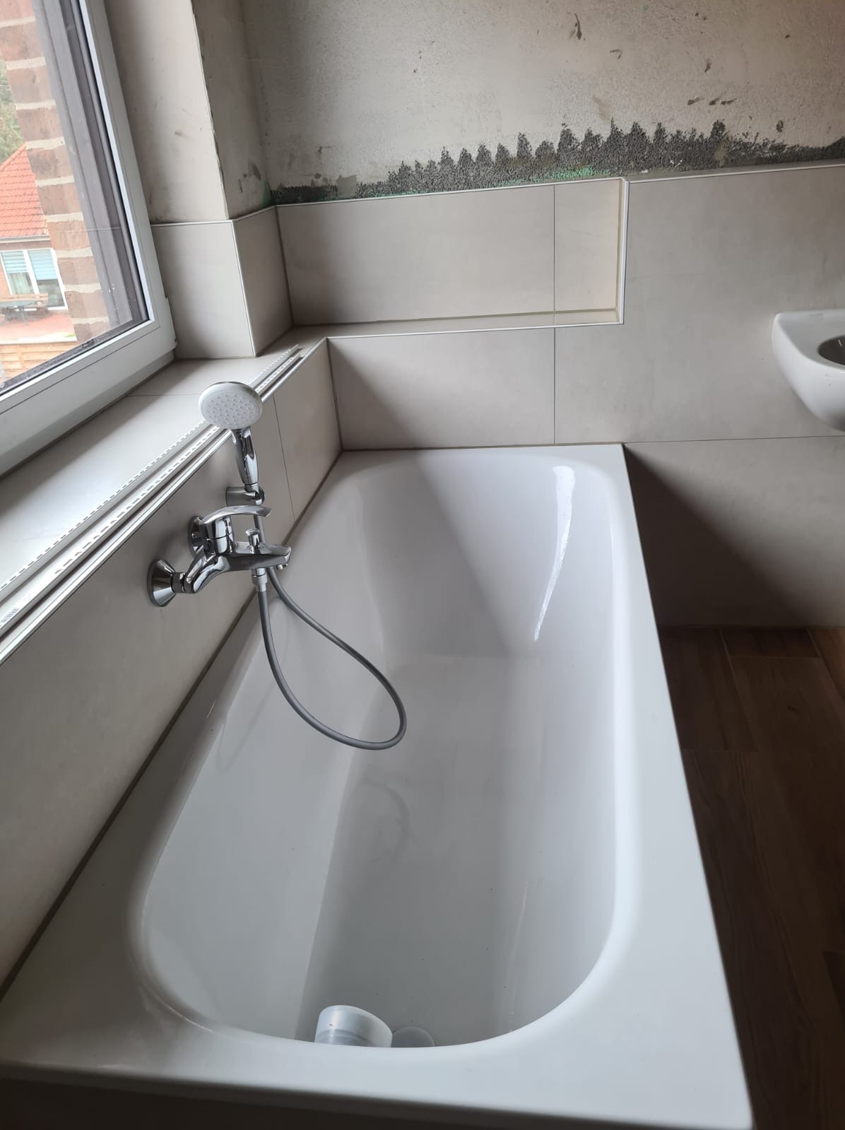 Biała wanna w trakcie montażu, widoczna armatura, fragment ściany z położonymi płytkami ceramicznymi i surową powierzchnią powyżej, okno z widokiem na budynek z czerwonej cegły.