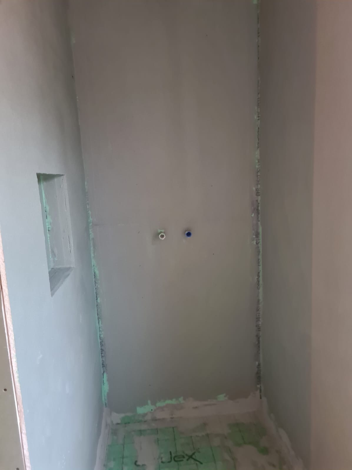 Przygotowana do wykończenia ściana w łazience z wnęką i wyprowadzonymi przyłączami wodnymi, widoczna izolacja podłogowa z zielonym nadrukiem.