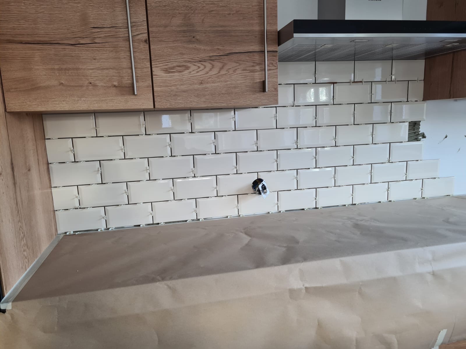 Układanie białych, prostokątnych płytek ceramicznych typu metro na ścianie nad blatem kuchennym, widoczne krzyżyki dystansowe i przygotowane gniazdko elektryczne, zabezpieczony blat roboczy.