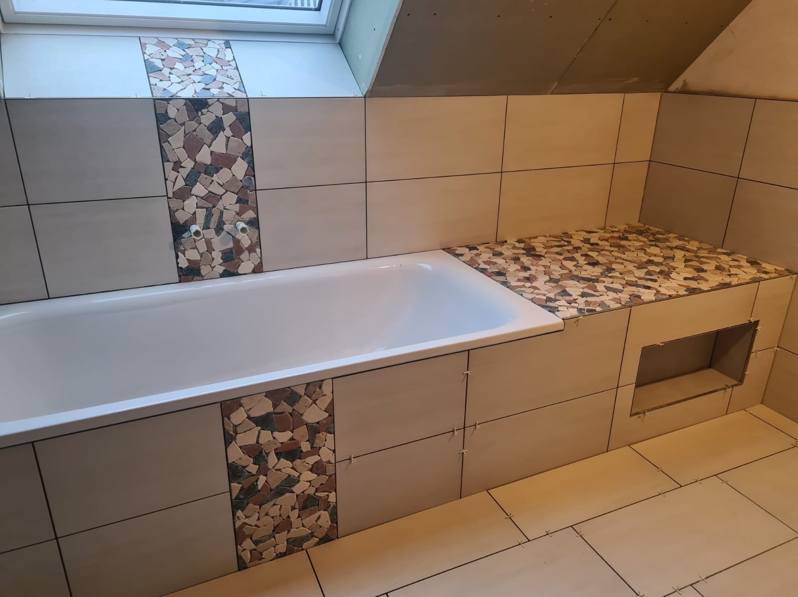 Łazienka w trakcie wykańczania: biała wanna obudowana płytkami w odcieniach beżu, z dekoracyjnymi pasami i fragmentami z mozaiki w kolorach ziemi. Widoczne krzyżyki dystansowe między płytkami.