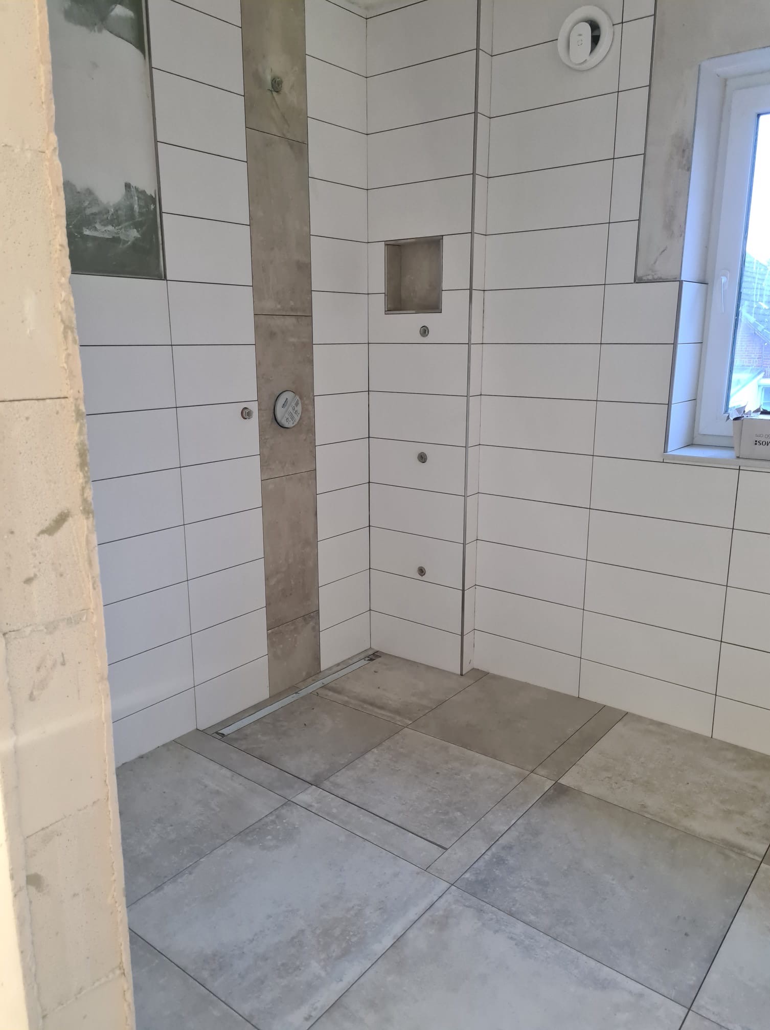 Surowa łazienka w trakcie prac: białe, prostokątne płytki ścienne z ciemnymi fugami, szara kolumna z instalacją, szare płytki podłogowe, widoczny otwór wentylacyjny i okno.