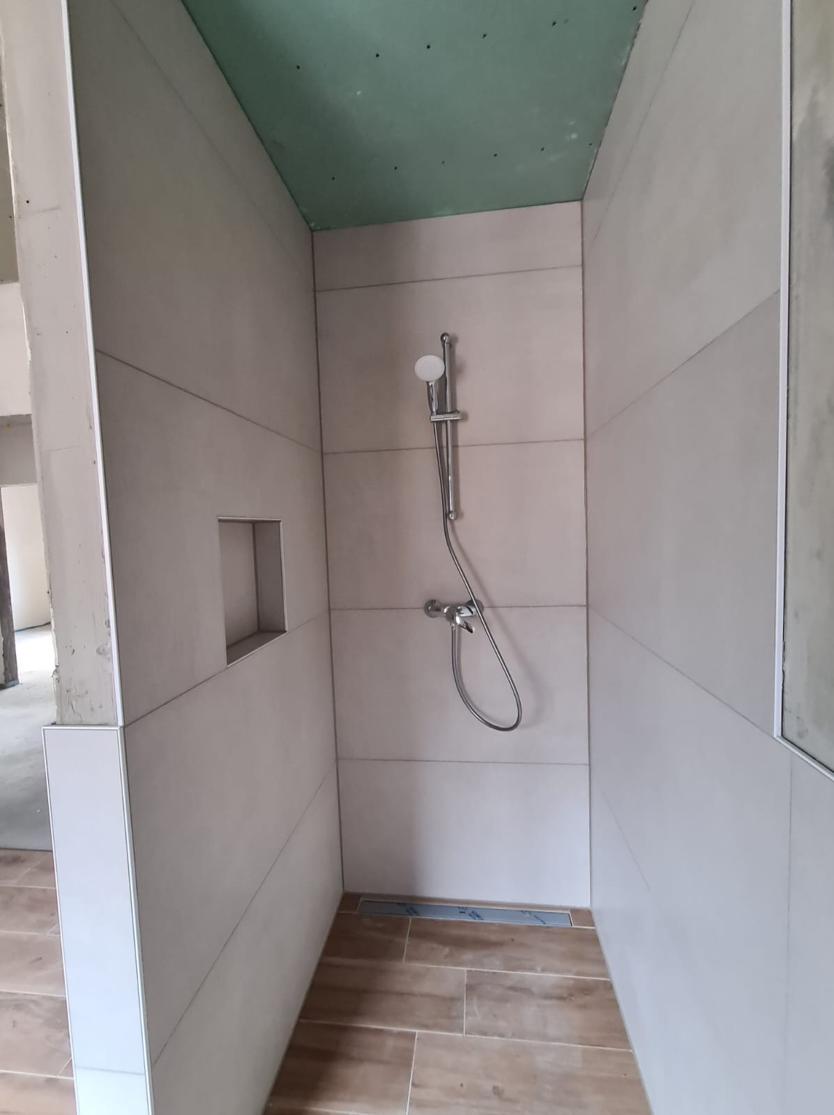 Płytki ścienne w odcieniach beżu ułożone poziomo w kabinie prysznicowej z wnęką w ścianie, widoczna armatura i odpływ liniowy w podłodze z płytek imitujących drewno, sufit wykończony płytą...