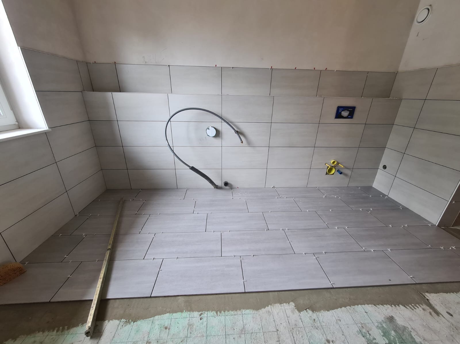 Układanie szarych płytek w łazience na podłodze i ścianach, widoczne krzyżyki dystansowe i wystające rury instalacyjne, w tle okno.