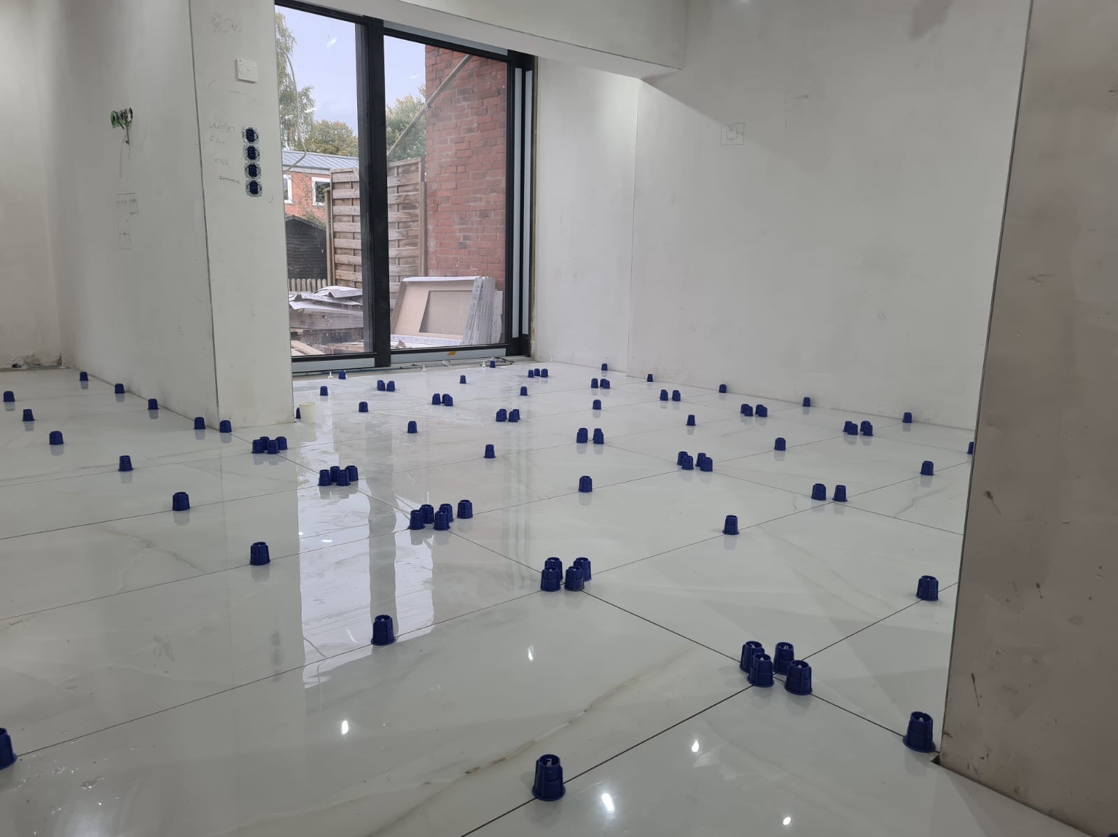 Białe, błyszczące płytki podłogowe z niebieskimi elementami poziomującymi, ułożone w pustym pomieszczeniu z dużym oknem i instalacją elektryczną na ścianie.