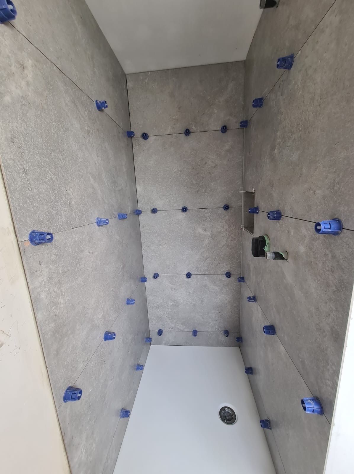 Szare płytki imitujące kamień na ścianach i suficie łazienki, wyrównywane za pomocą niebieskich klipsów i sznurków, widoczny biały brodzik z odpływem.
