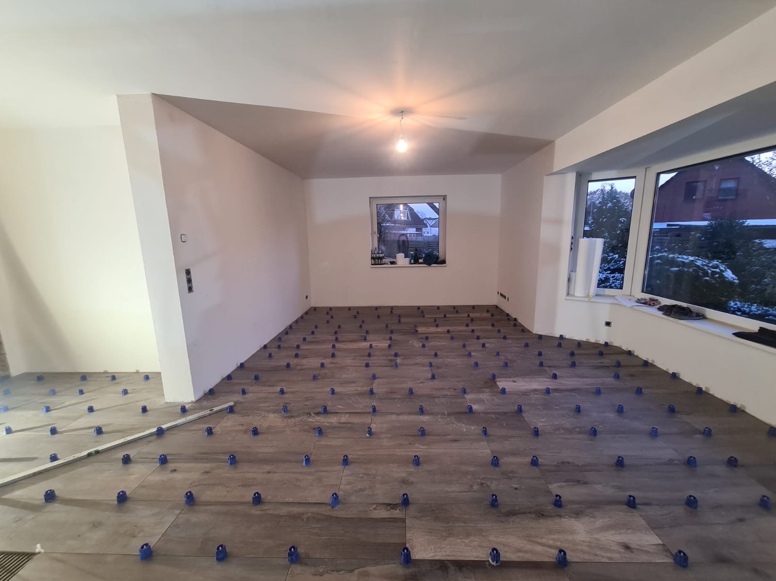 Układanie płytek podłogowych imitujących drewno z użyciem niebieskich klipsów poziomujących w przestronnym, jasnym pokoju podczas remontu.