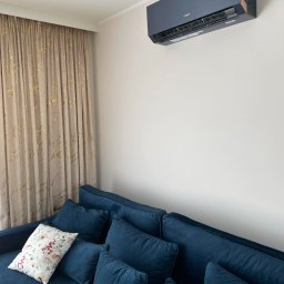 MK KLIMA MICHAŁ KUBECZKA - Ciemnoszary klimatyzator zamontowany na ścianie w pokoju nad granatową sofą z poduszkami, obok okna z zasłoną w odcieniach beżu i złota.