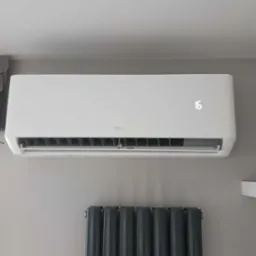 Biały klimatyzator TCL zamontowany na szarej ścianie, wyświetlający temperaturę 16 stopni Celsjusza, poniżej grzejnik panelowy w kolorze grafitowym.