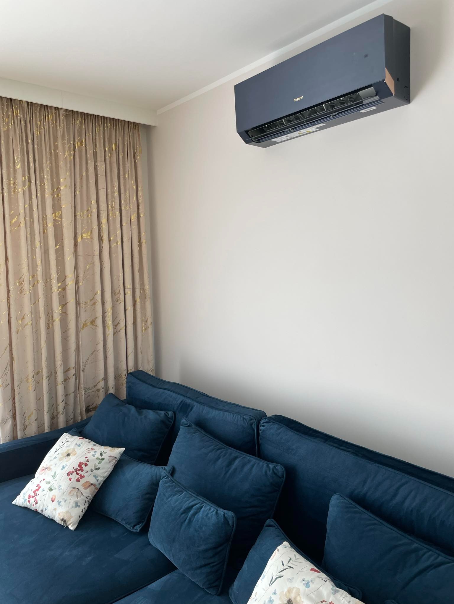 Ciemnoszary klimatyzator zamontowany na ścianie w pokoju nad granatową sofą z poduszkami, obok okna z zasłoną w odcieniach beżu i złota.