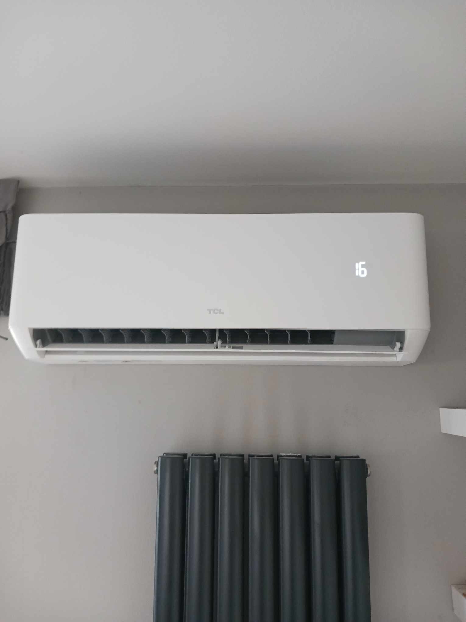 Biały klimatyzator TCL zamontowany na szarej ścianie, wyświetlający temperaturę 16 stopni Celsjusza, poniżej grzejnik panelowy w kolorze grafitowym.
