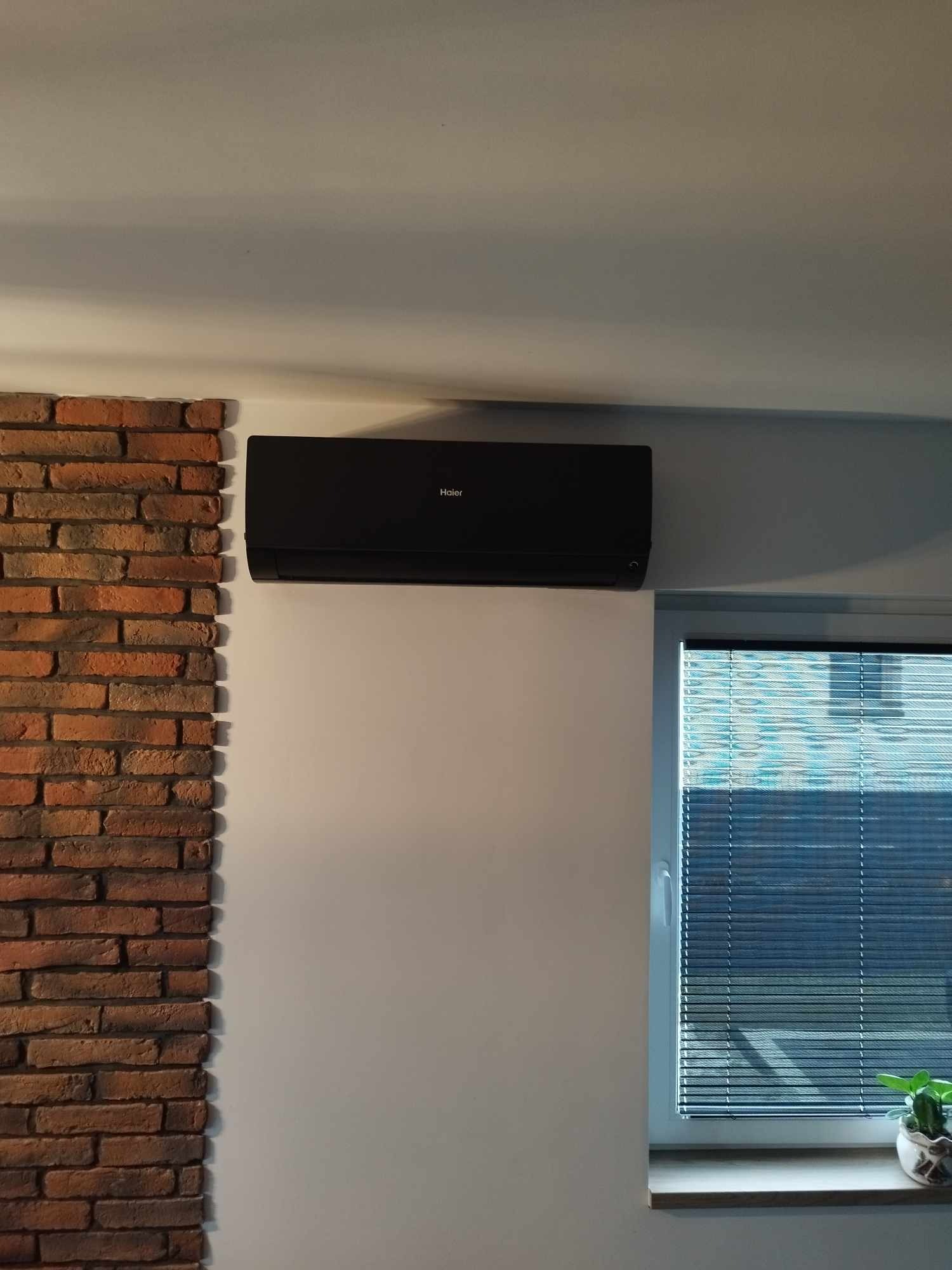 Czarna jednostka klimatyzacyjna Haier zamontowana na białej ścianie w pomieszczeniu z ceglaną dekoracją i oknem z żaluzjami.