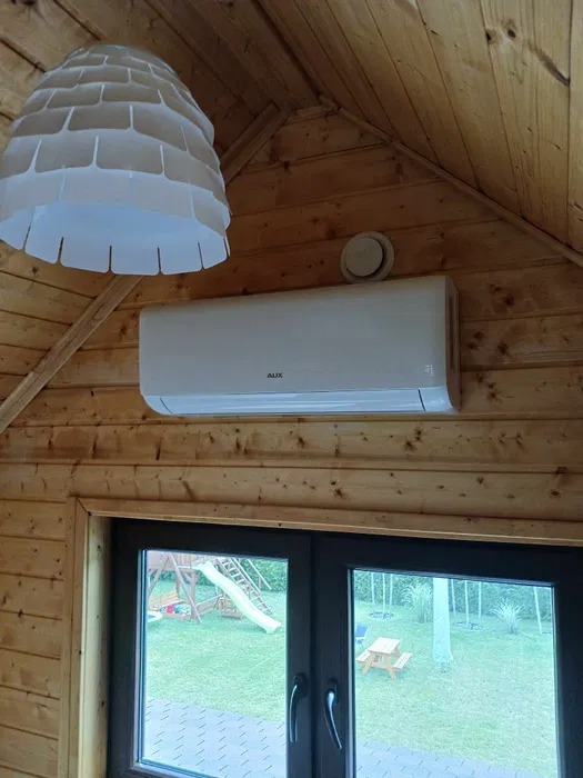 Biały klimatyzator AUX zamontowany na drewnianej ścianie w domku letniskowym, widok z okna na plac zabaw.