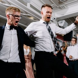 Moose Wedding Fotografia - Rozbawieni goście weselni tańczą i śpiewają z otwartymi ustami, widoczna para młoda w tle, oświetlenie parkietu.