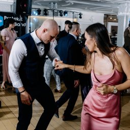 Moose Wedding Fotografia - Para w różowych strojach tańczy na parkiecie podczas imprezy, w tle widoczni inni goście i sprzęt nagłośnieniowy.