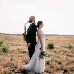 Para młoda stoi plecami do siebie na tle suchej trawy; mężczyzna trzyma saksofon.