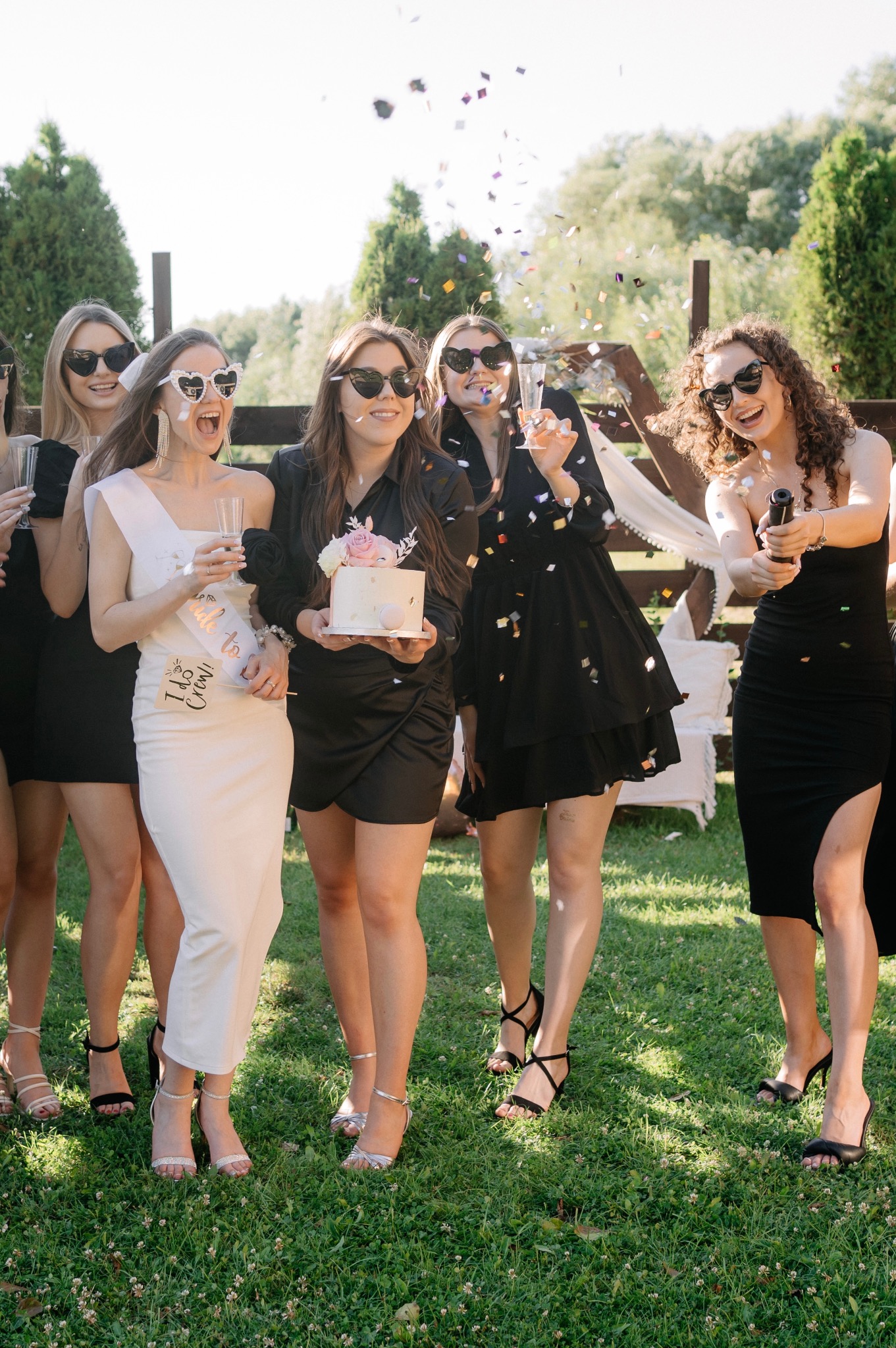 Grupa kobiet świętuje na zewnątrz, konfetti w powietrzu, jedna trzyma tort z różowymi kwiatami, inne w okularach w kształcie serc i z szampanem.