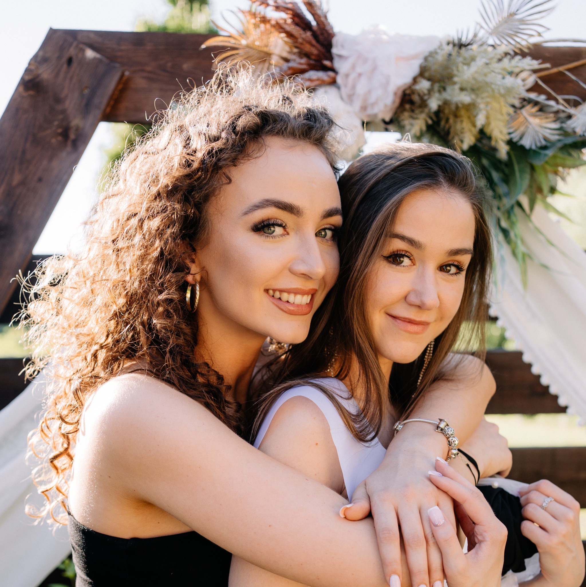 Dwie uśmiechnięte kobiety obejmują się na tle drewnianej konstrukcji udekorowanej kwiatami i białym materiałem, prawdopodobnie podczas sesji zdjęciowej.