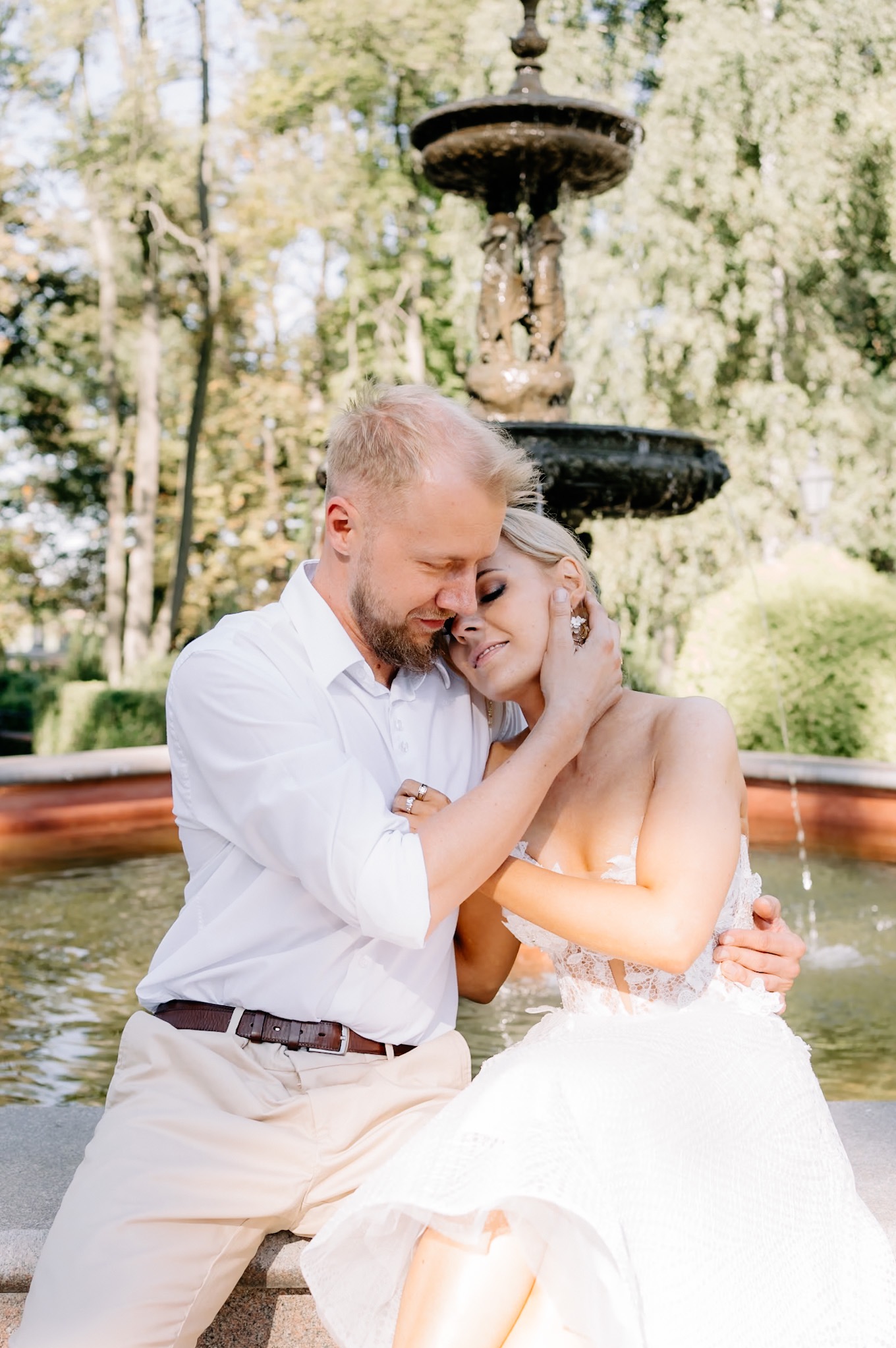 Para młoda w romantycznym uścisku na tle fontanny w Ostrowcu Świętokrzyskim, panna młoda w koronkowej sukni z odsłoniętymi ramionami, pan młody w białej koszuli i beżowych spodniach.