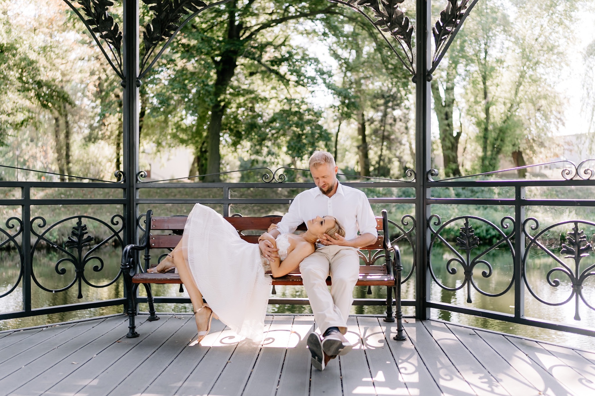 Para młoda w altanie nad wodą: panna młoda w koronkowej sukni leży na kolanach pana młodego, który siedzi na ławce, w tle drzewa i woda.
