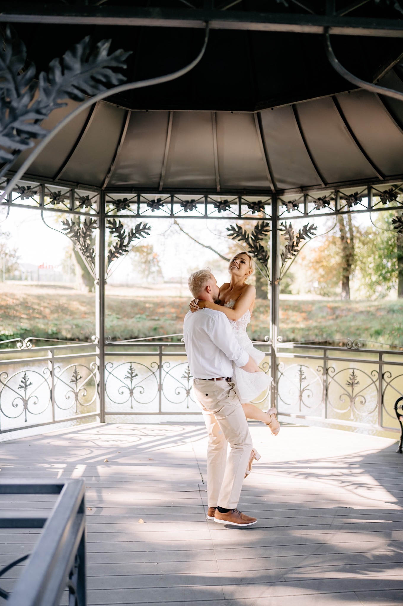 Uśmiechnięta panna młoda w białej sukni podnoszona przez pana młodego w jasnych spodniach i białej koszuli w ozdobnej altanie z widokiem na park.